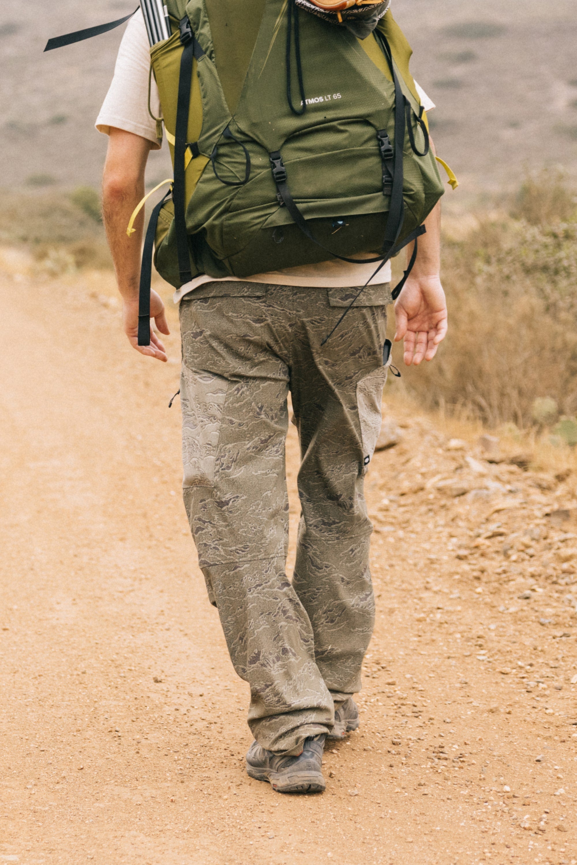 Wearing a large green backpack and 686 Men's Anything Cargo Pant - Relaxed Fit, a person walks along a dirt trail in a dry, natural landscape, seen from behind.