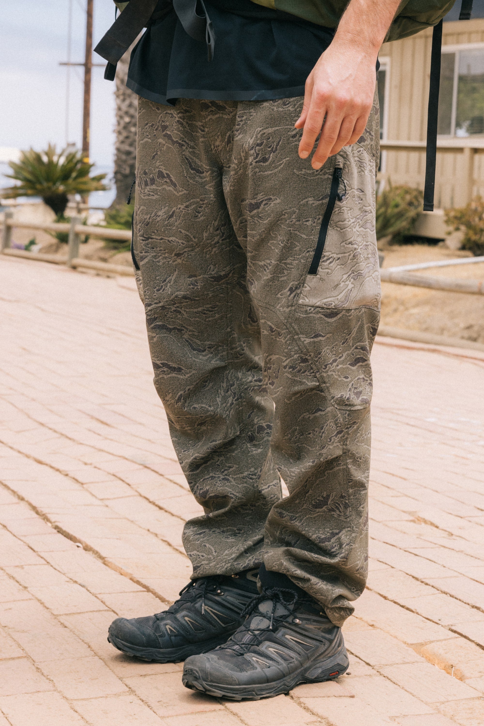 A person outdoors, visible from the waist down, wears the 686 Men's Anything Cargo Pant - Relaxed Fit with 4-way stretch and zipper pockets, paired with black trail shoes.
