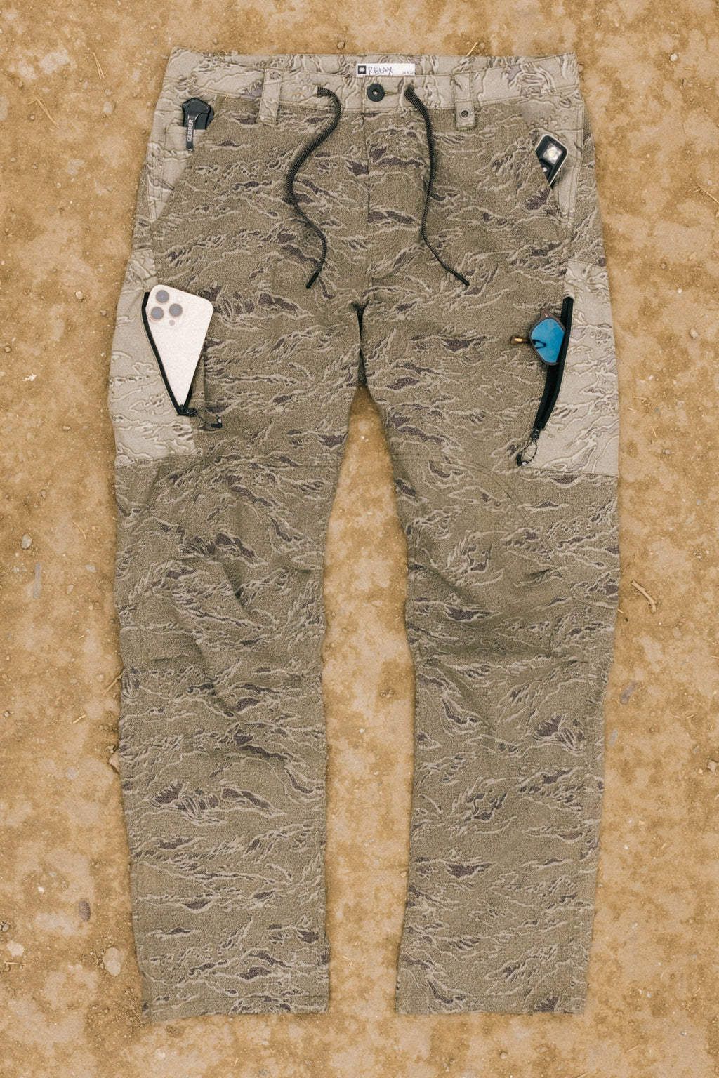 The 686 Men's Anything Cargo Pant - Relaxed Fit from 686, featuring a camouflage pattern and 13 pockets, rests on dirt with a phone and utility tool in the pockets and drawstrings visible at the waist.