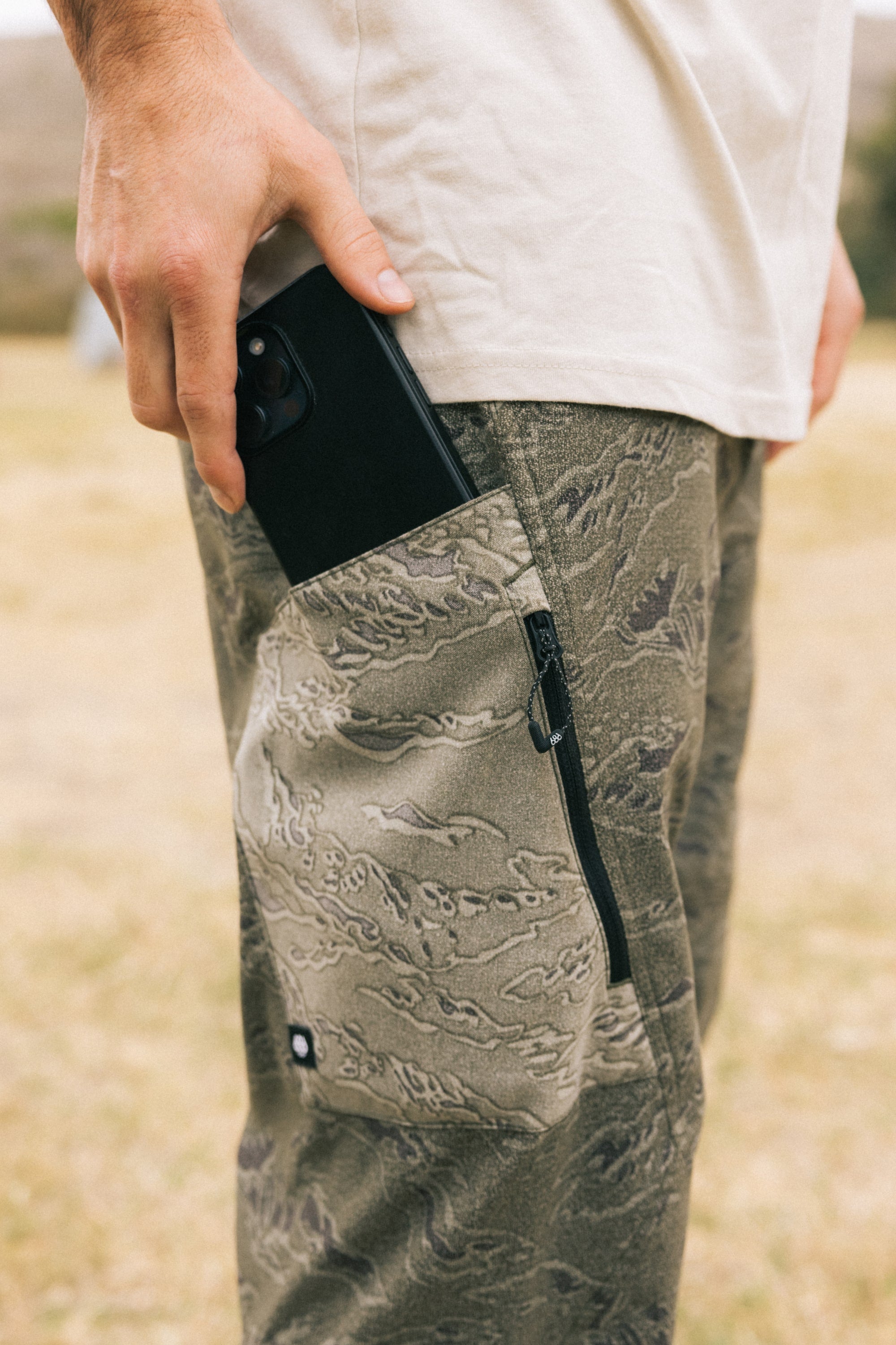 A person puts a black smartphone into the zippered side pocket of their 686 Men's Anything Cargo Pant - Relaxed Fit by 686 while standing outdoors.
