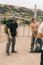 Two men stand on a dirt path by a metal gate and dry hills. One wears a hat, sunglasses, and a backpack; the other is shirtless in 686 Men's Anything Cargo Pant - Relaxed Fit by 686, sunglasses, and holding a backpack.