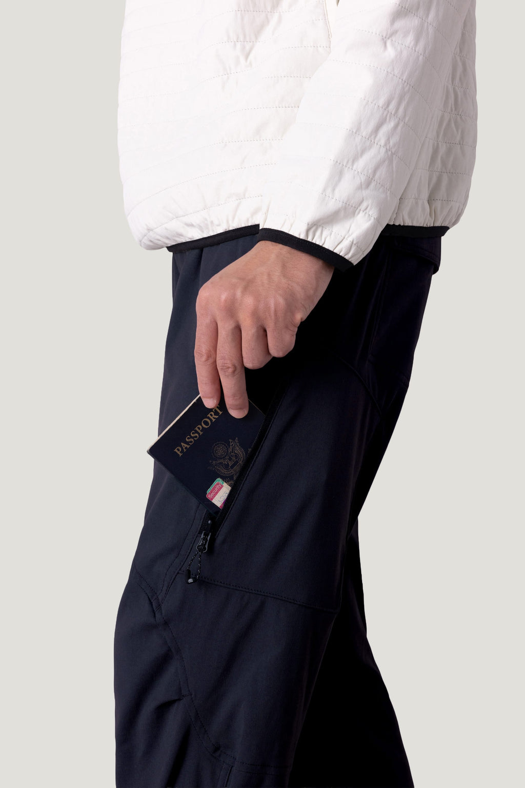 A person in a white jacket and dark pants holds a dark passport partially tucked into the zippered pocket of their 686 Men's Anything Cargo Pant - Relaxed Fit by 686.