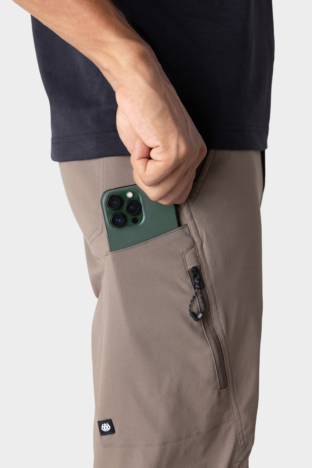 Wearing a black shirt and 686 Men's Anything Cargo Pant - Relaxed Fit in taupe, a person slips a green smartphone into the open thigh pocket of these technical cargo pants featuring 4-way stretch and zippered pockets.