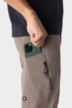 Wearing a black shirt and 686 Men's Anything Cargo Pant - Relaxed Fit in taupe, a person slips a green smartphone into the open thigh pocket of these technical cargo pants featuring 4-way stretch and zippered pockets.