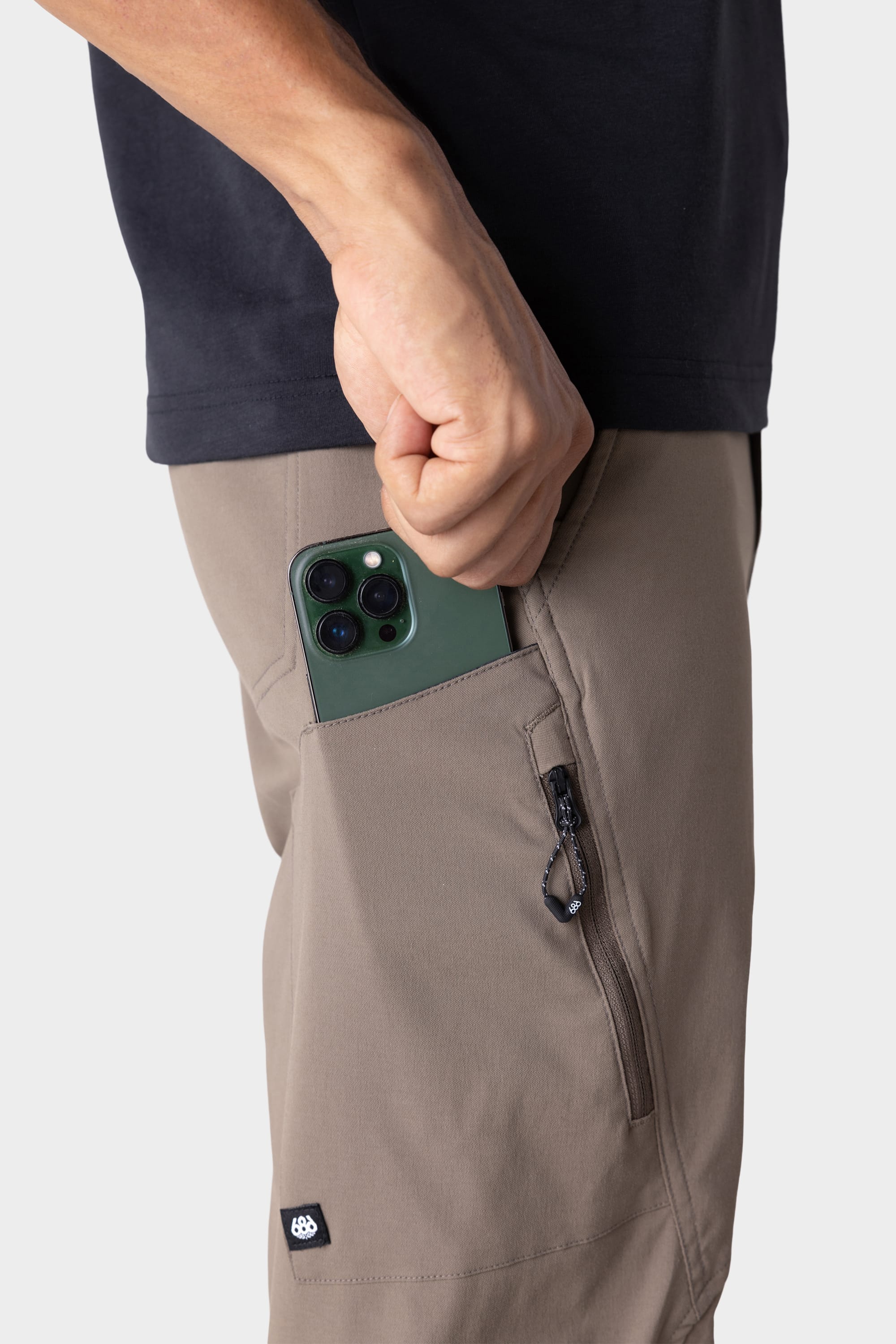 Wearing a black shirt and 686 Men's Anything Cargo Pant - Relaxed Fit in taupe, a person slips a green smartphone into the open thigh pocket of these technical cargo pants featuring 4-way stretch and zippered pockets.