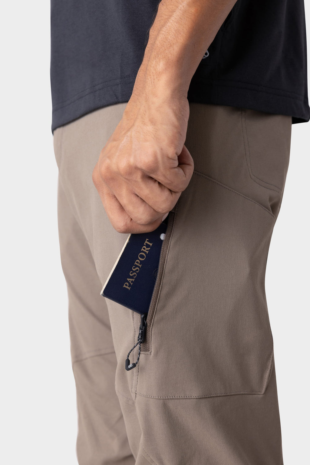 A person’s hand holds a navy blue passport, partially tucked into the zippered pocket of the 686 Men's Anything Cargo Pant - Relaxed Fit in light brown. Only their lower torso and hand, along with a dark shirt, are visible.
