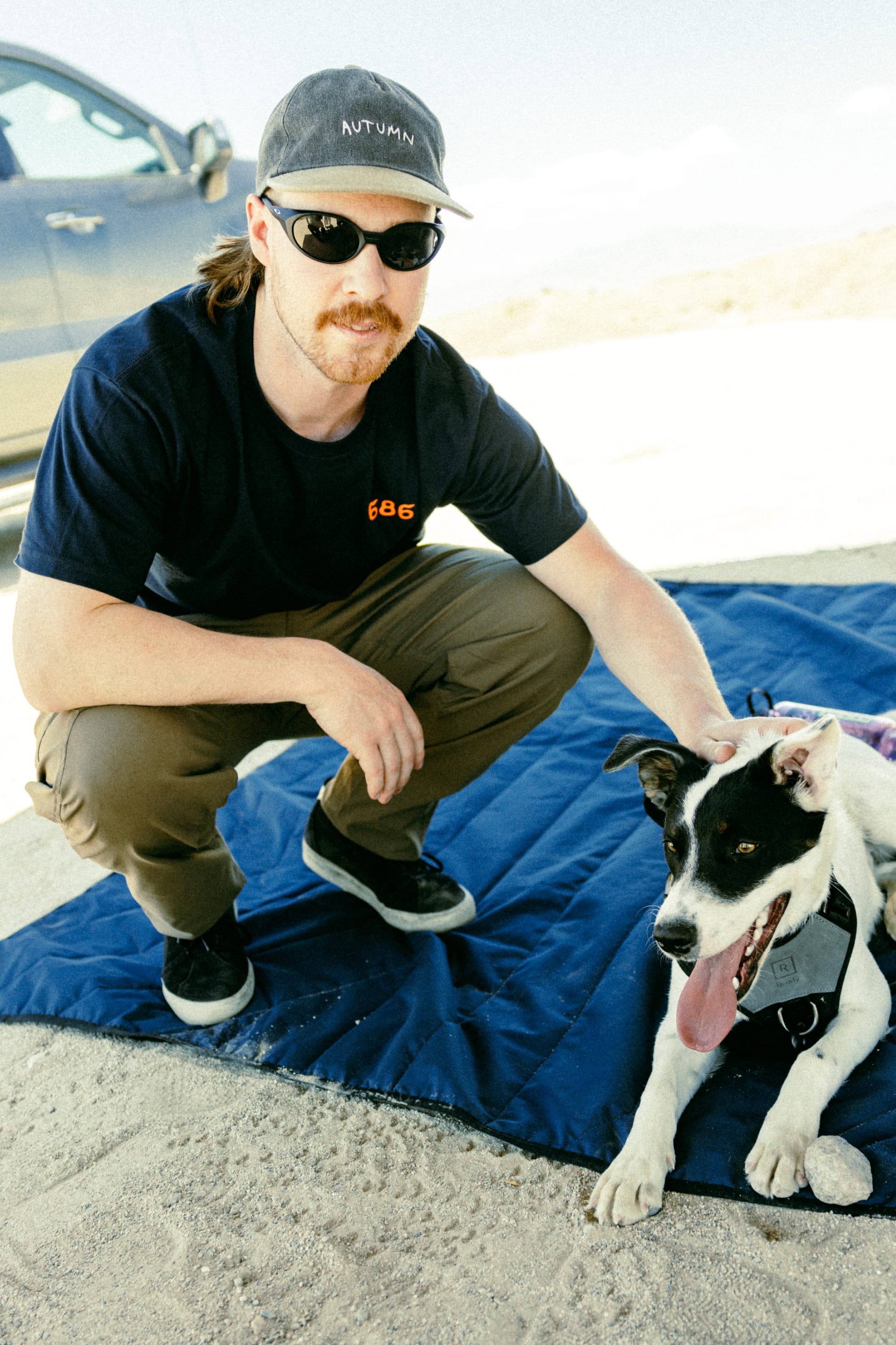 A man in sunglasses, a cap, and dark T-shirt kneels on a blue mat outdoors, petting a black-and-white dog. He's wearing 686 Men's Anything Cargo Pant - Relaxed Fit by 686. A car and sandy ground are visible in the background.
