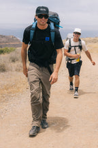 Two men in hats, sunglasses, and backpacks hike a dry coastal trail. The man in front smiles as the other carries a drink—both wearing 686 Men's Anything Cargo Pant - Relaxed Fit, designed by 686 for comfort and easy movement.