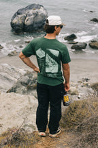 Wearing a green graphic tee, white cap, sunglasses, and holding a drink bottle, a person stands on a rocky ocean cliff with waves behind, sporting 686 Men's Everywhere® Pant - Relaxed Fit by 686.