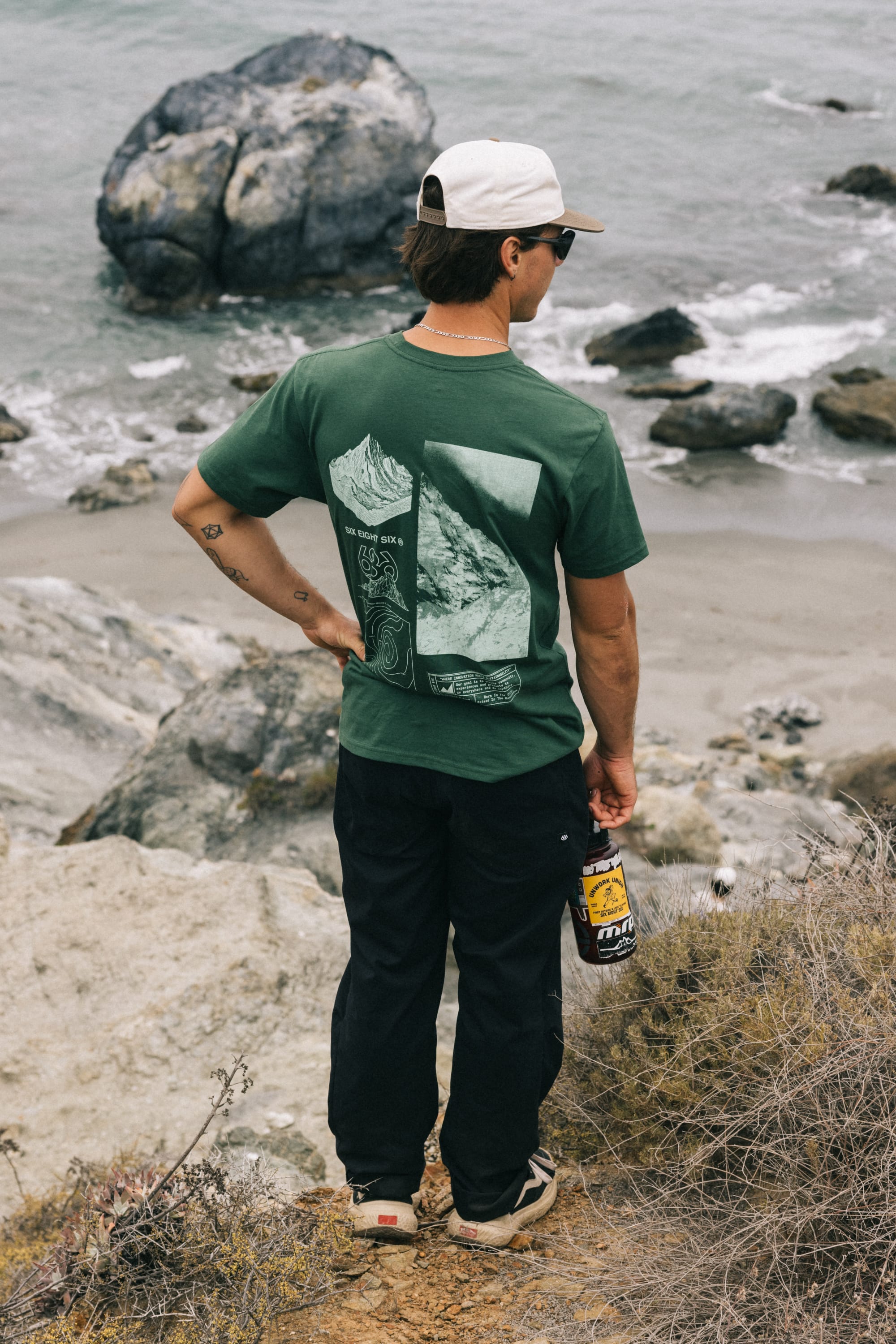 Wearing a green graphic tee, white cap, sunglasses, and holding a drink bottle, a person stands on a rocky ocean cliff with waves behind, sporting 686 Men's Everywhere® Pant - Relaxed Fit by 686.