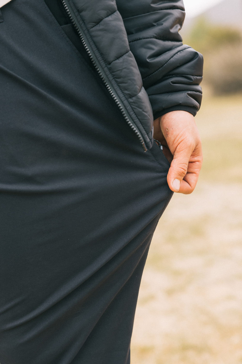 A man outdoors in a black jacket and 686 Men's Everywhere® Pant - Relaxed Fit pulls at the pant fabric with his right hand; blurred grass and greenery form the background.