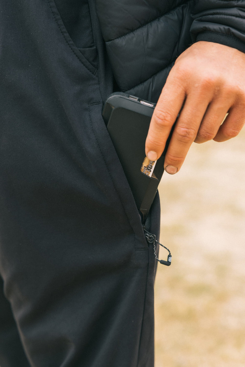 A hand places a black smartphone into the zippered side pocket of 686 Men's Everywhere® Pant - Relaxed Fit, with only the hand, pocket, and part of a black jacket visible. Brand: 686.
