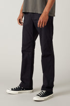 A person wearing the 686 Men's Everywhere® Pant - Relaxed Fit in a dark shade, paired with a gray shirt and black low-top sneakers with white soles, stands against a neutral background, shown from the lower chest down.