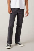 Only the lower half of a person is visible, wearing a white t-shirt, 686 Men's Everywhere® Pant - Relaxed Fit in dark gray, and black Converse sneakers against a light gray background.