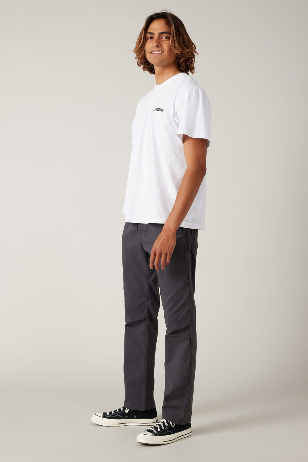 A young man with medium-length brown hair stands sideways, smiling. He wears a white t-shirt, 686 Men's Everywhere® Pant - Relaxed Fit by 686, and black sneakers with white soles, against a plain light background.
