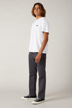 A young man with medium-length brown hair stands sideways, smiling. He wears a white t-shirt, 686 Men's Everywhere® Pant - Relaxed Fit by 686, and black sneakers with white soles, against a plain light background.