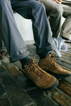 Two people sit comfortably on a wooden floor, only their legs and feet visible. One wears brown hiking boots with gray breathable pants; the other sports gray sneakers with 686 Men's Everywhere® Pant - Relaxed Fit by 686 in light khaki.