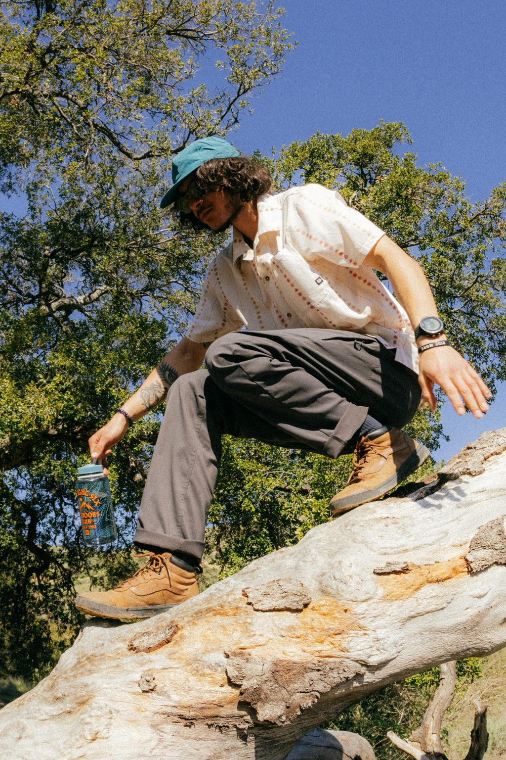 A person wearing 686 Men's Everywhere® Pant - Relaxed Fit by 686, a blue cap, sunglasses, patterned shirt, and hiking boots climbs over a fallen tree trunk in the forest, holding a water bottle amid green trees under a clear sky.