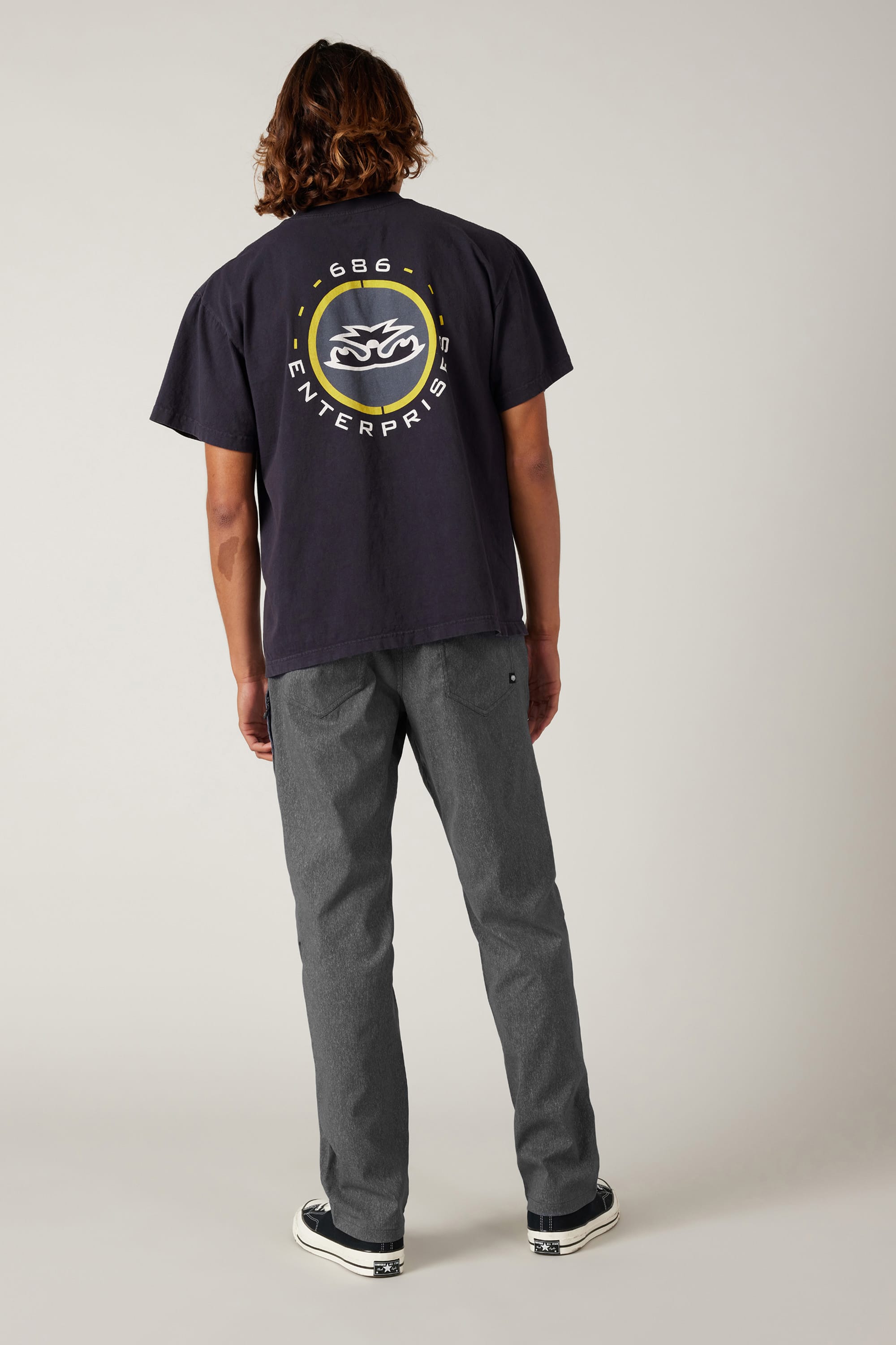 A person with shoulder-length hair stands facing away, wearing a black T-shirt with a large circular logo, 686 Men's Everywhere® Pant - Relaxed Fit from 686, and black sneakers, against a plain light background.