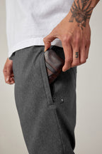 A person in a white shirt and 686 Men's Everywhere® Pant - Relaxed Fit slides a brown leather wallet into their pocket, revealing tattoos on their hand and forearm.