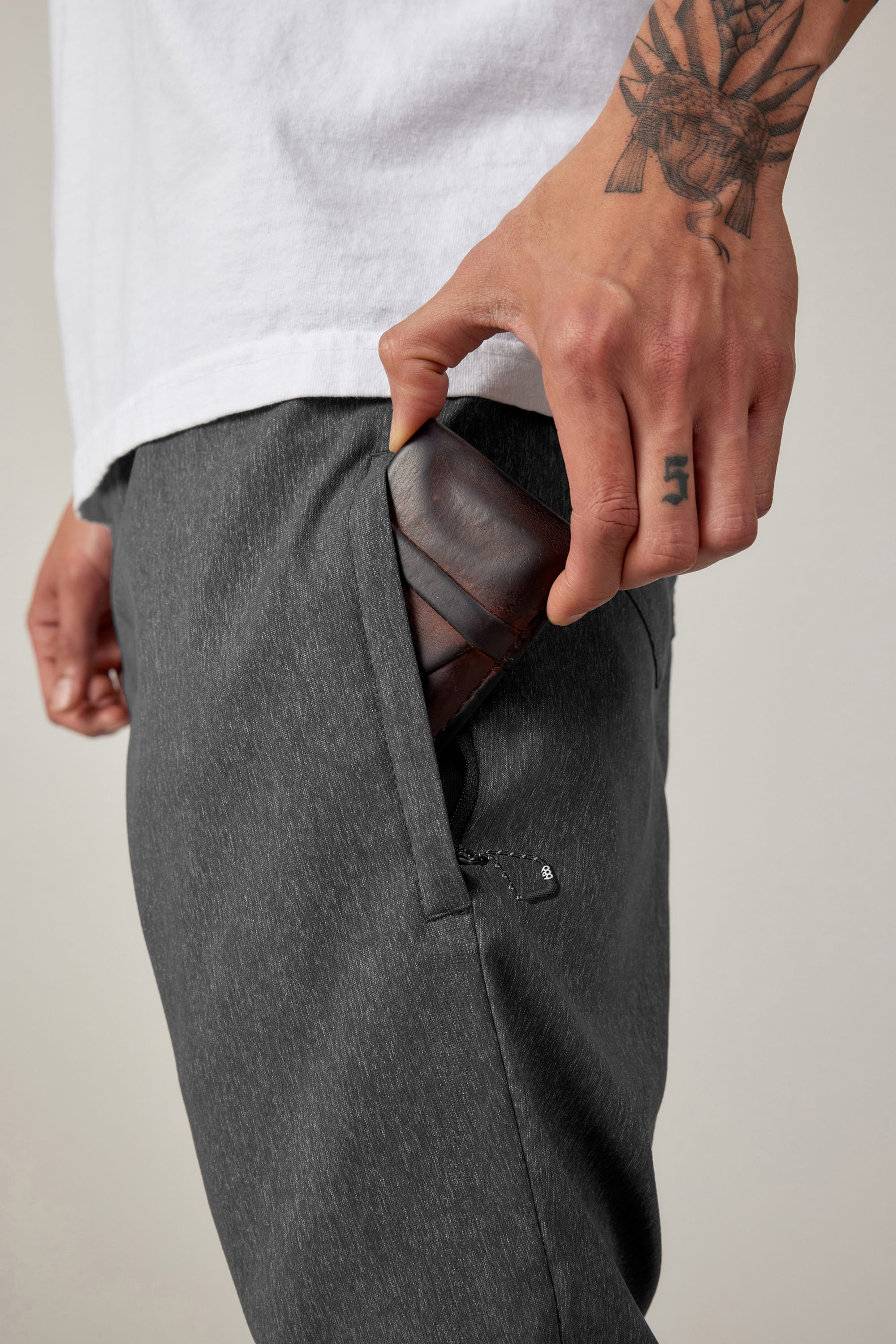 A person in a white shirt and 686 Men's Everywhere® Pant - Relaxed Fit slides a brown leather wallet into their pocket, revealing tattoos on their hand and forearm.