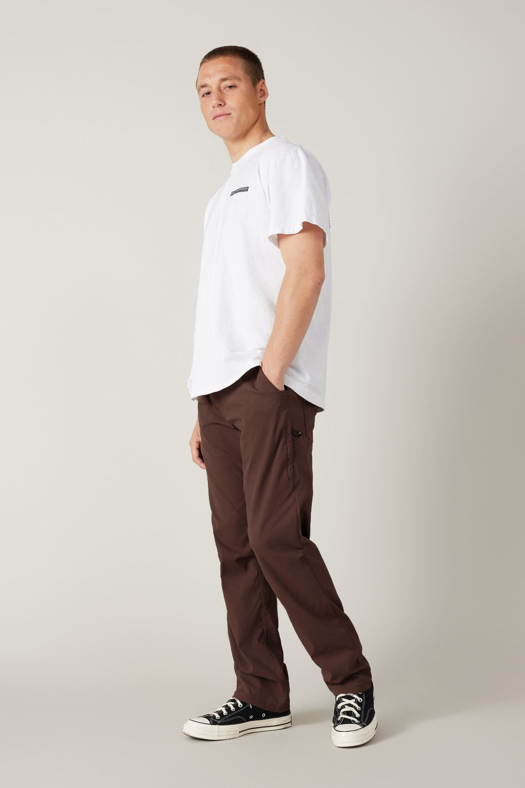 A person stands in a relaxed pose against a plain backdrop, wearing a white t-shirt, 686 Men's Everywhere® Pant - Relaxed Fit in brown, and black-and-white sneakers. Their left hand is in their pocket as they look at the camera.