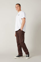 A person stands in a relaxed pose against a plain backdrop, wearing a white t-shirt, 686 Men's Everywhere® Pant - Relaxed Fit in brown, and black-and-white sneakers. Their left hand is in their pocket as they look at the camera.