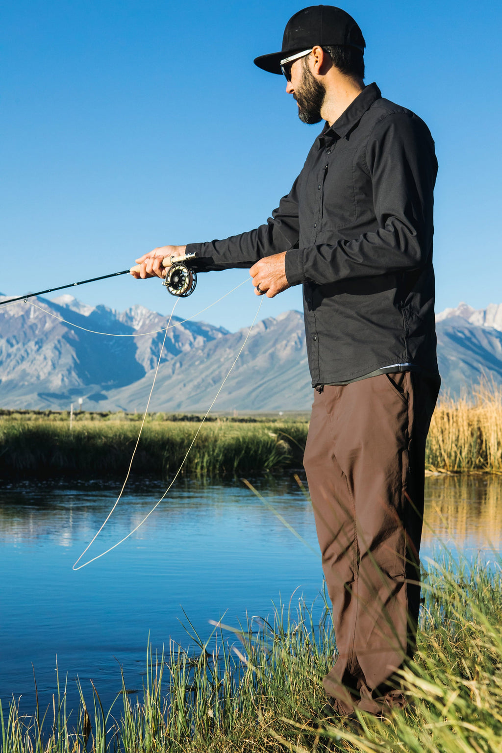 A man wearing a black shirt, 686 Men's Everywhere® Pant - Relaxed Fit, and a cap is fly fishing by a river with mountains and a clear blue sky in the background.