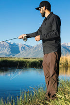 A man wearing a black shirt, 686 Men's Everywhere® Pant - Relaxed Fit, and a cap is fly fishing by a river with mountains and a clear blue sky in the background.