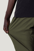 A person wearing 686 Men's Everywhere® Pant - Relaxed Fit in olive green stretches the flexible fabric with one hand, while their arm and part of a black shirt are also visible.