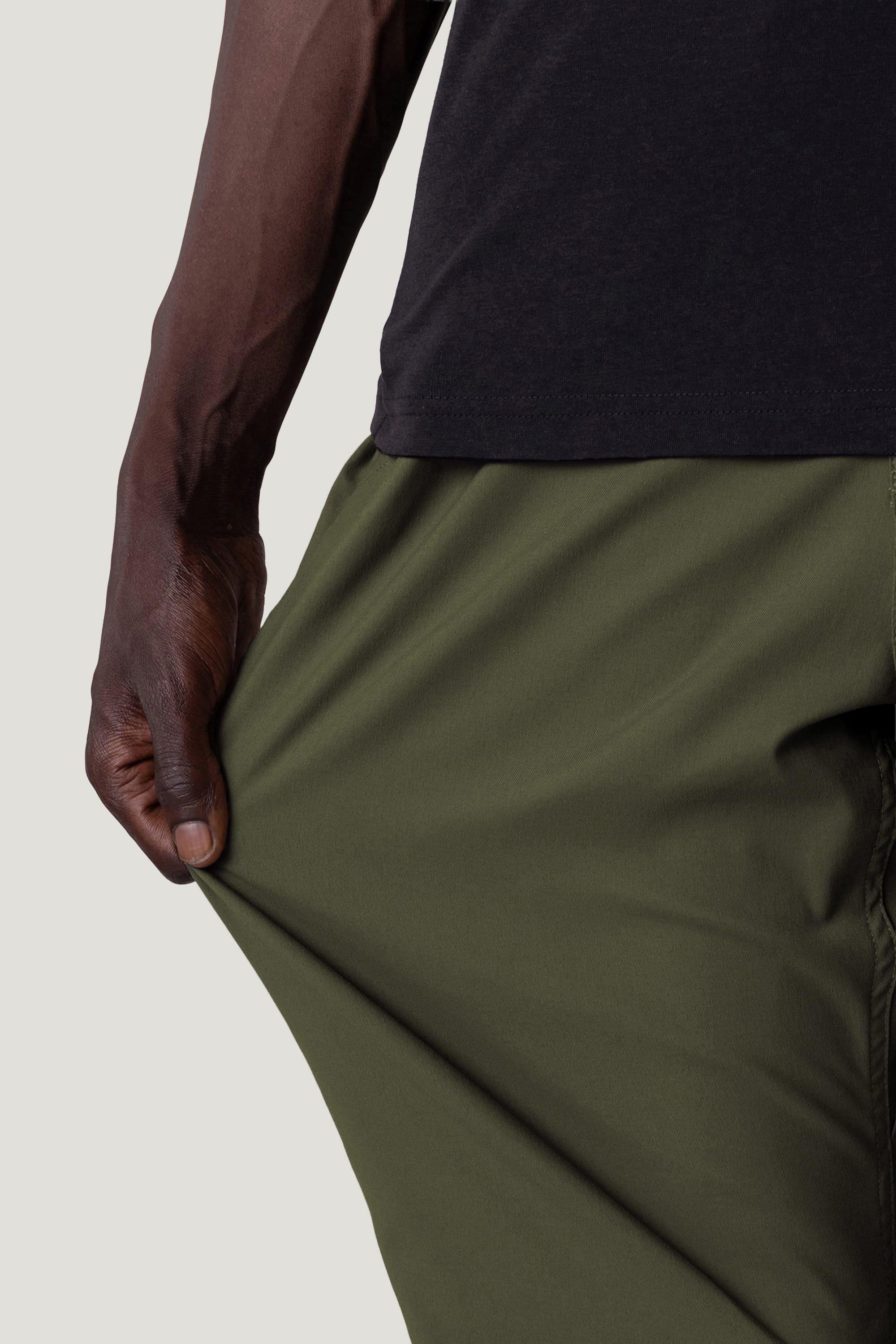A person wearing 686 Men's Everywhere® Pant - Relaxed Fit in olive green stretches the flexible fabric with one hand, while their arm and part of a black shirt are also visible.