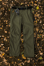The 686 Men's Everywhere® Pant - Relaxed Fit in olive green, featuring a black belt and climbing gear on the front belt loop, is displayed flat on soil scattered with yellow and brown leaves.