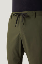 A close-up shows a person wearing 686 Men's Everywhere® Pant - Relaxed Fit in olive green, featuring a black drawstring waist and paired with a dark tucked-in shirt. Only the waist and upper thigh are visible.