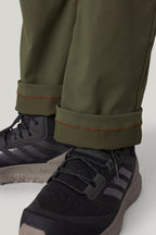 Close-up of a person wearing the 686 Men's Everywhere® Pant - Relaxed Fit in dark green with rolled cuffs and orange stitching, paired with black athletic shoes featuring gray stripes and thick soles.
