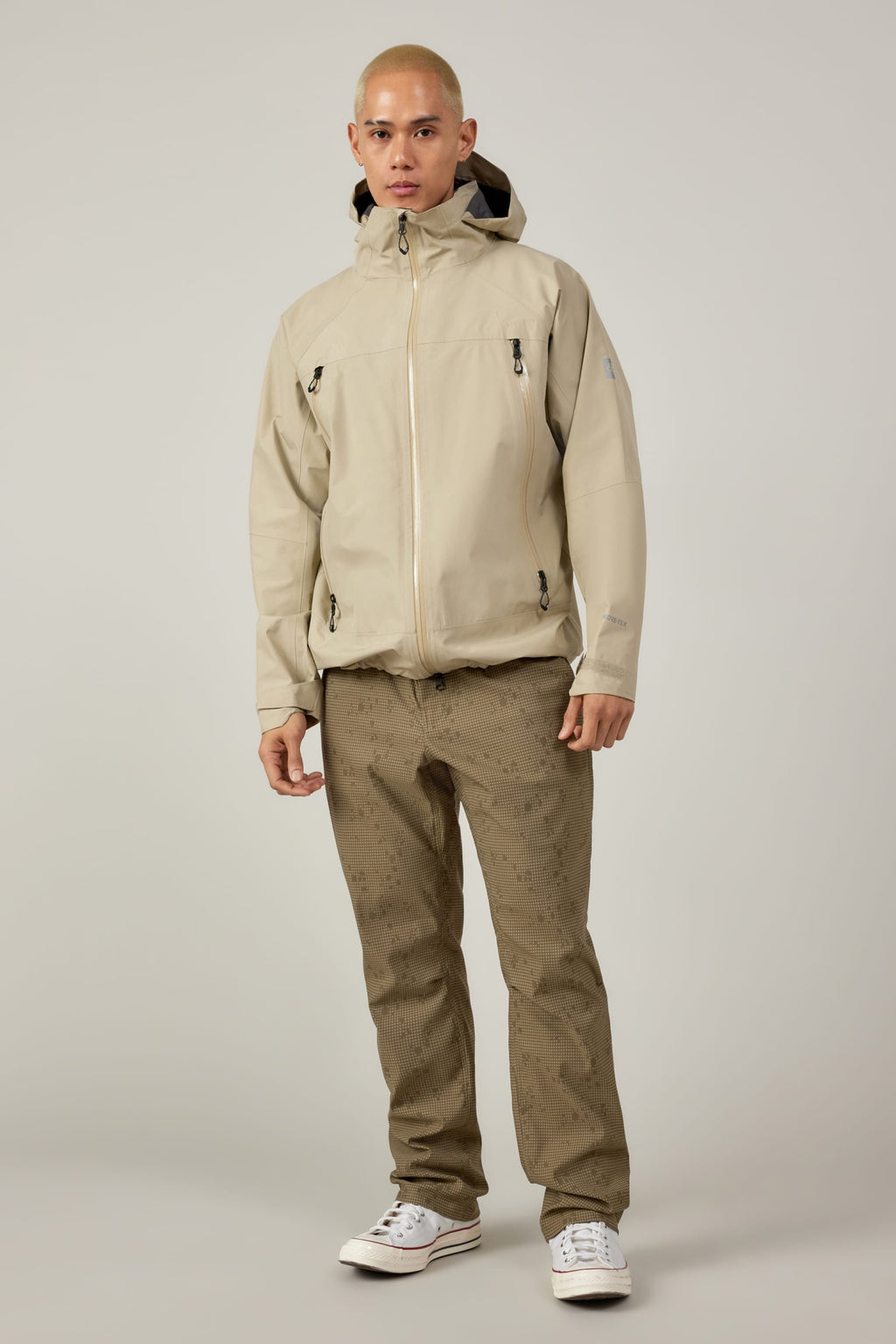 A person with a shaved head wears a light beige hooded jacket, 686 Men's Everywhere® Pant - Relaxed Fit in olive green with a water droplet pattern, and white sneakers, standing against a plain light gray background.