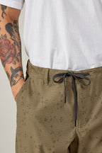 A person with colorful tattoos, including a red rose, wears a white t-shirt and 686 Men's Everywhere® Pant - Relaxed Fit in olive green, featuring a subtle dotted pattern and one hand in their pocket.