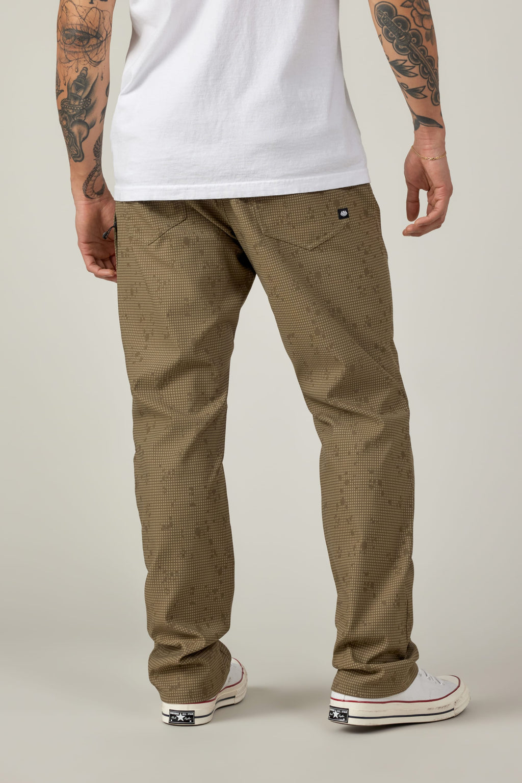 A person with tattooed arms models the 686 Men's Everywhere® Pant - Relaxed Fit in a tan check pattern, paired with a white T-shirt and white sneakers, standing facing away to show the back view against a plain background.