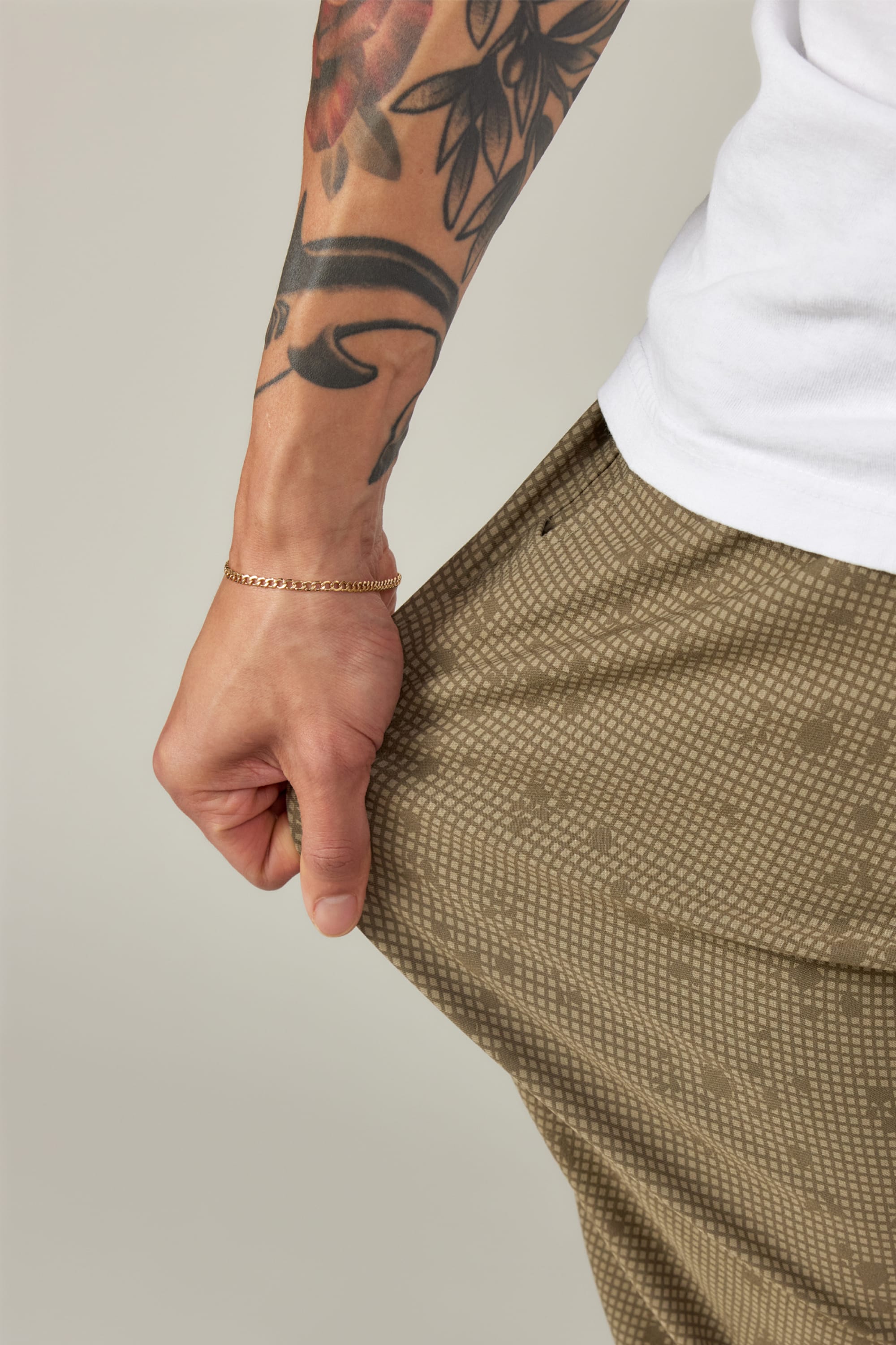 A person with a tattooed arm and gold chain bracelet pulls at the pocket of their brown 686 Men's Everywhere® Pant - Relaxed Fit. They wear a white shirt against a plain, neutral background.