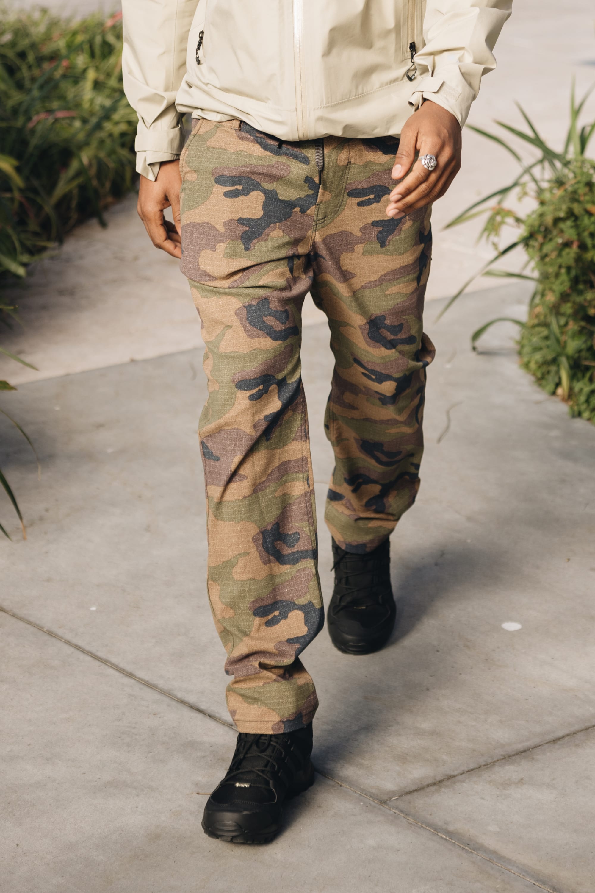 A person wearing 686 Men's Everywhere® Pant - Relaxed Fit and black shoes walks on a sidewalk lined with greenery. Only the lower half of the body is shown.