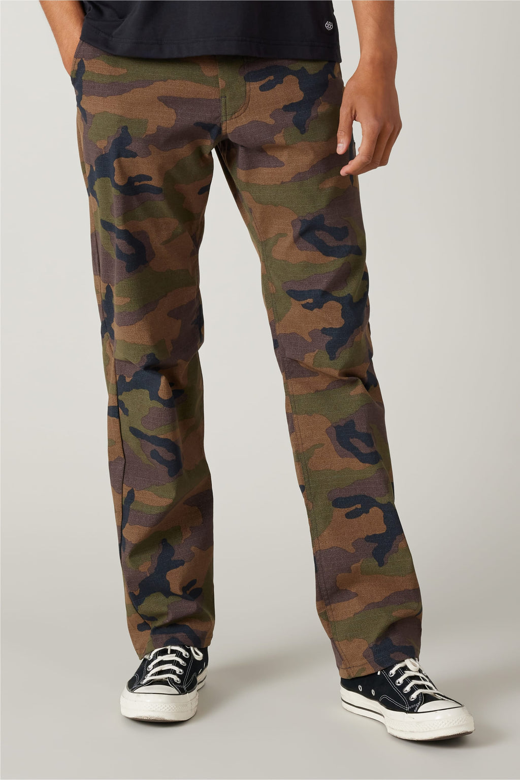 The image shows a person wearing 686 Men's Everywhere® Pant - Relaxed Fit in a camouflage pattern from 686, paired with a black T-shirt and black Converse sneakers, standing against a plain light background with only the lower body visible.