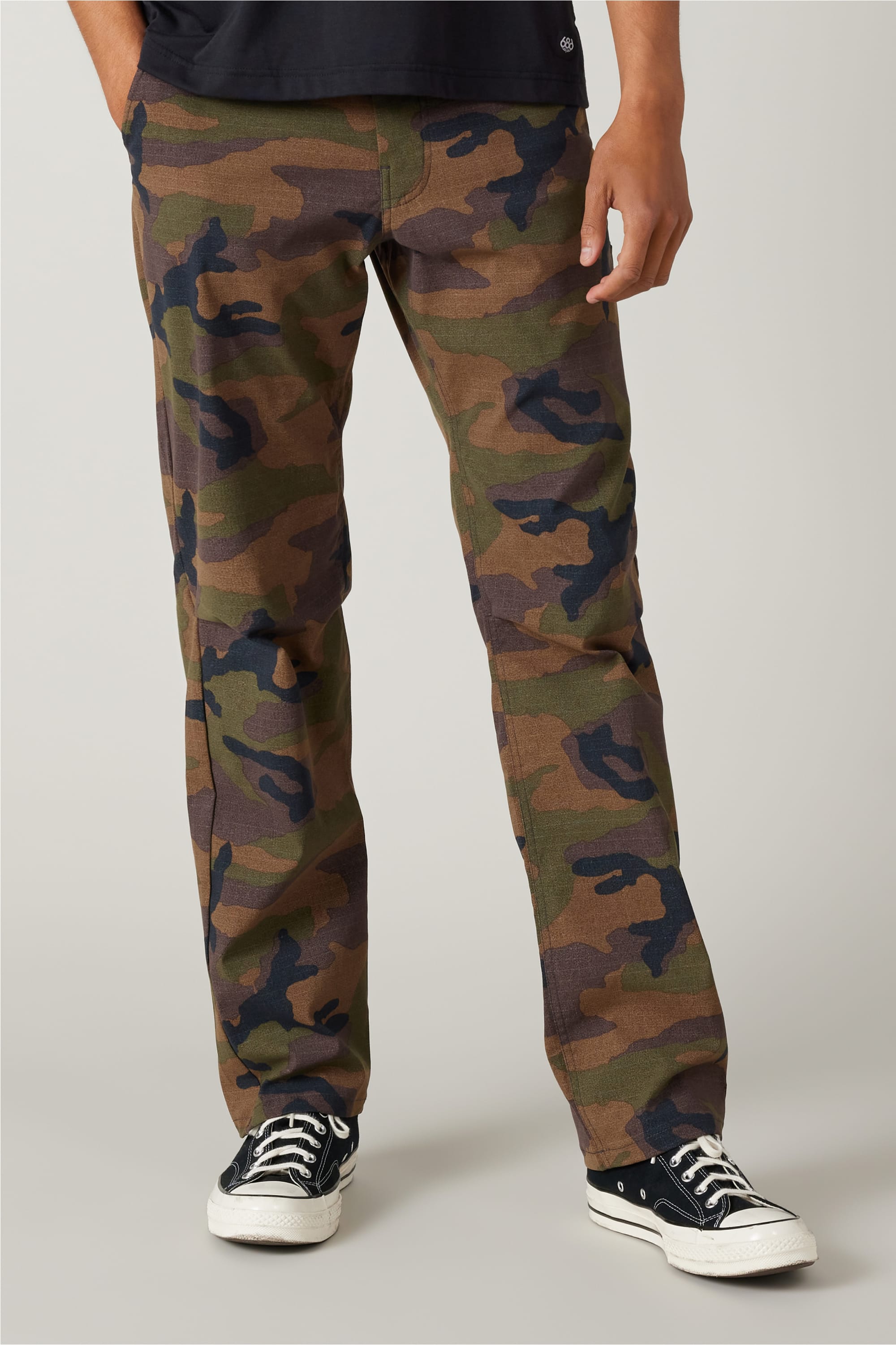The image shows a person wearing 686 Men's Everywhere® Pant - Relaxed Fit in a camouflage pattern from 686, paired with a black T-shirt and black Converse sneakers, standing against a plain light background with only the lower body visible.