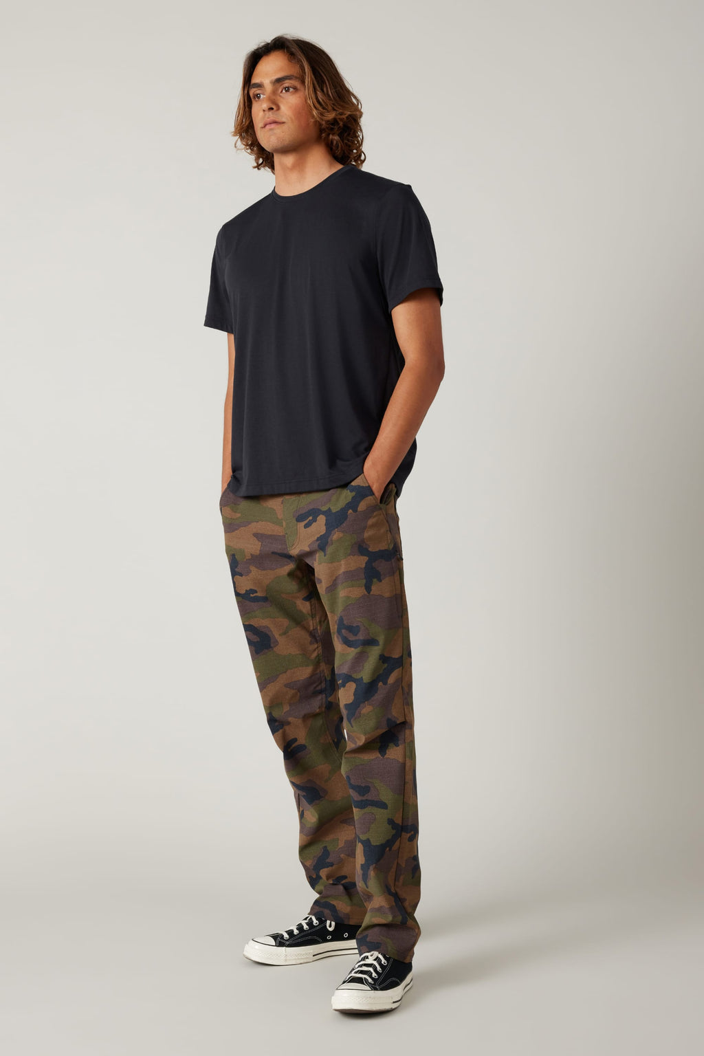 A person stands at an angle, wearing a plain black t-shirt, 686 Men's Everywhere® Pant - Relaxed Fit by 686 in camouflage, and black-and-white sneakers, hands in pockets, against a neutral background.