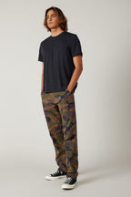 A person stands at an angle, wearing a plain black t-shirt, 686 Men's Everywhere® Pant - Relaxed Fit by 686 in camouflage, and black-and-white sneakers, hands in pockets, against a neutral background.