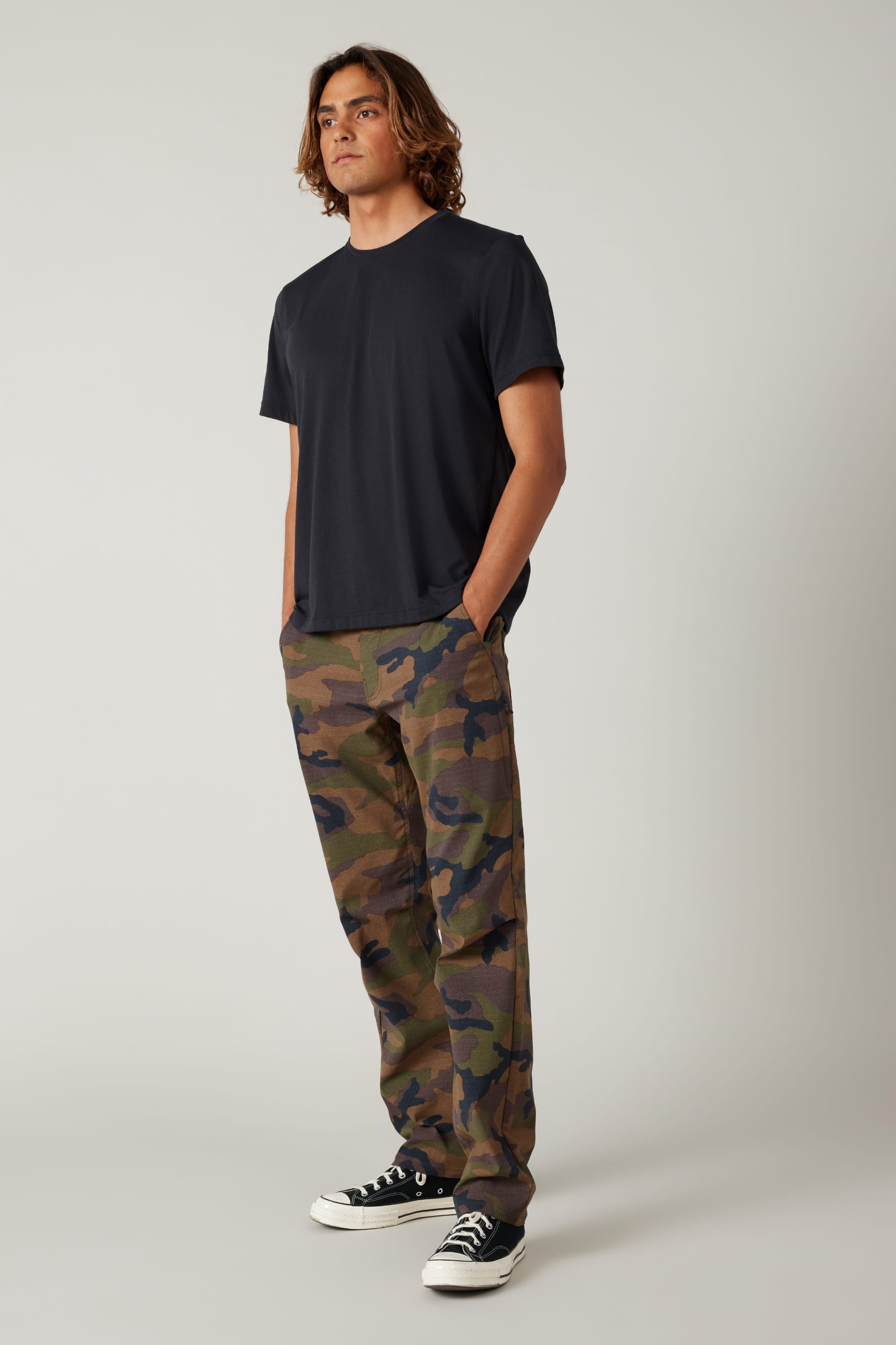 A person stands at an angle, wearing a plain black t-shirt, 686 Men's Everywhere® Pant - Relaxed Fit by 686 in camouflage, and black-and-white sneakers, hands in pockets, against a neutral background.