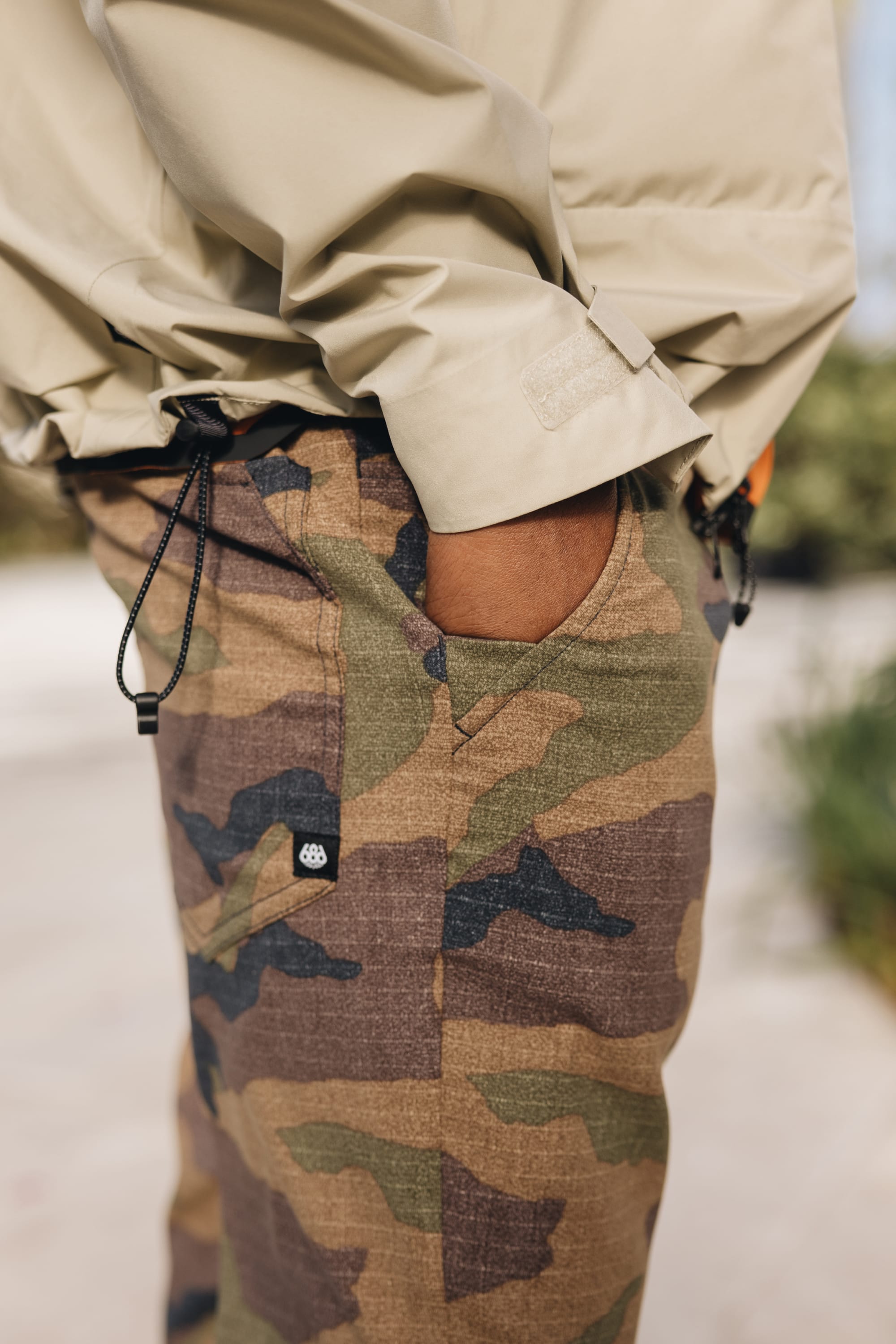 A person stands outdoors with one hand in their pocket, wearing a beige jacket and 686 Men's Everywhere® Pant - Relaxed Fit in camo. The breathable pants feature drawstrings and a small logo tag, highlighting the versatile 686 design.