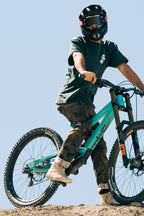 A person in a helmet, dark green t-shirt, and 686 Men's Everywhere® Pant - Relaxed Fit by 686 stands with one foot on the pedal of a teal mountain bike on dirt, with a clear blue sky in the background.