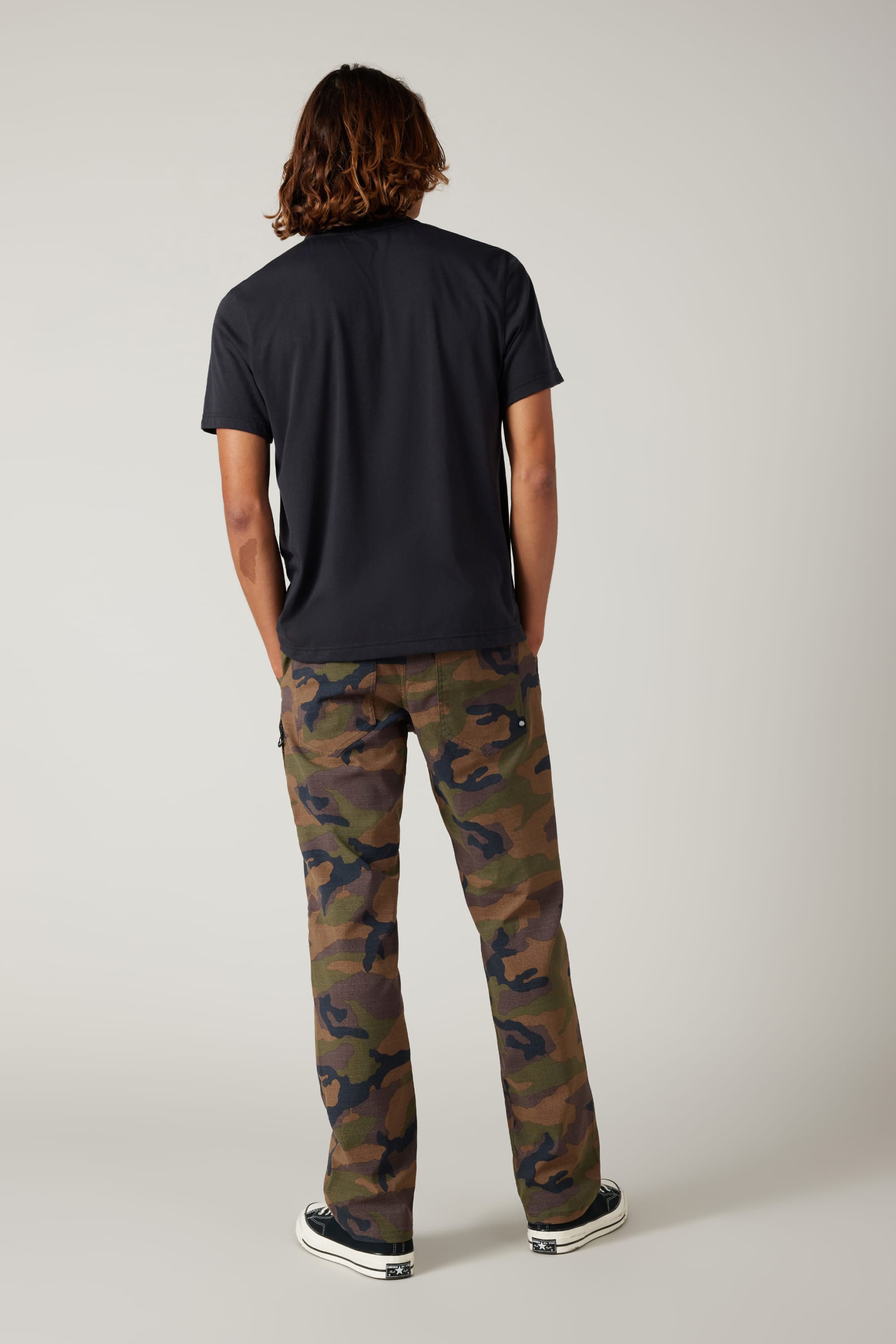 A person stands facing away against a plain light background, wearing a black t-shirt, 686 Men's Everywhere® Pant - Relaxed Fit by 686 in camouflage print, and black-and-white sneakers.