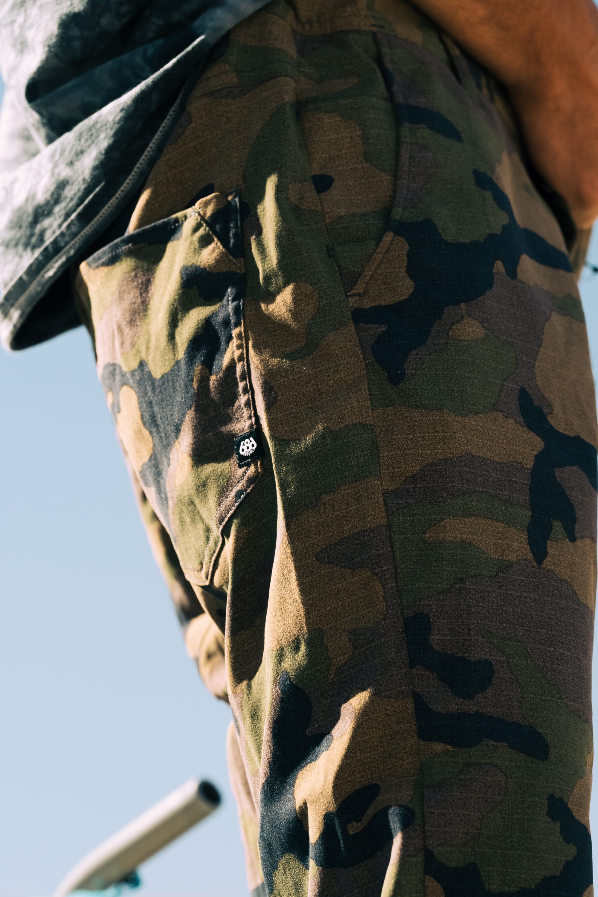 Close-up of a person wearing the 686 Men's Everywhere® Pant - Relaxed Fit by 686. Their hand rests at their side, showing the pocket and small button. The background features an out-of-focus blue sky.