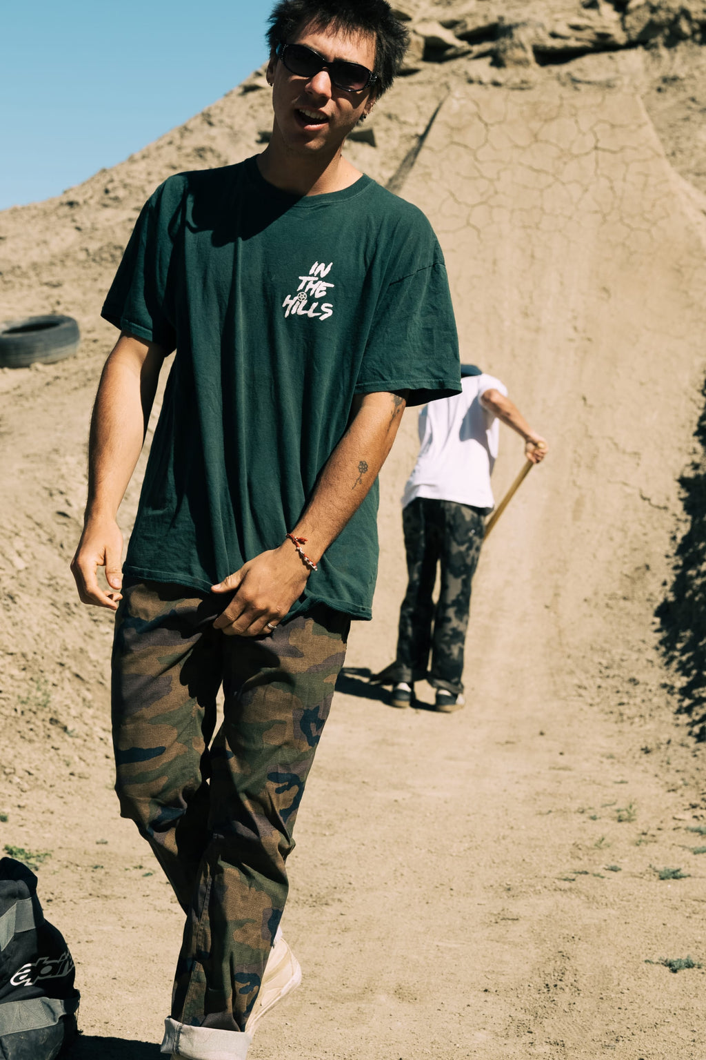 A young man in sunglasses and a dark green T-shirt stands on a dirt path wearing the 686 Men's Everywhere® Pant - Relaxed Fit, with another person holding a shovel in the background and sandy terrain surrounding them.