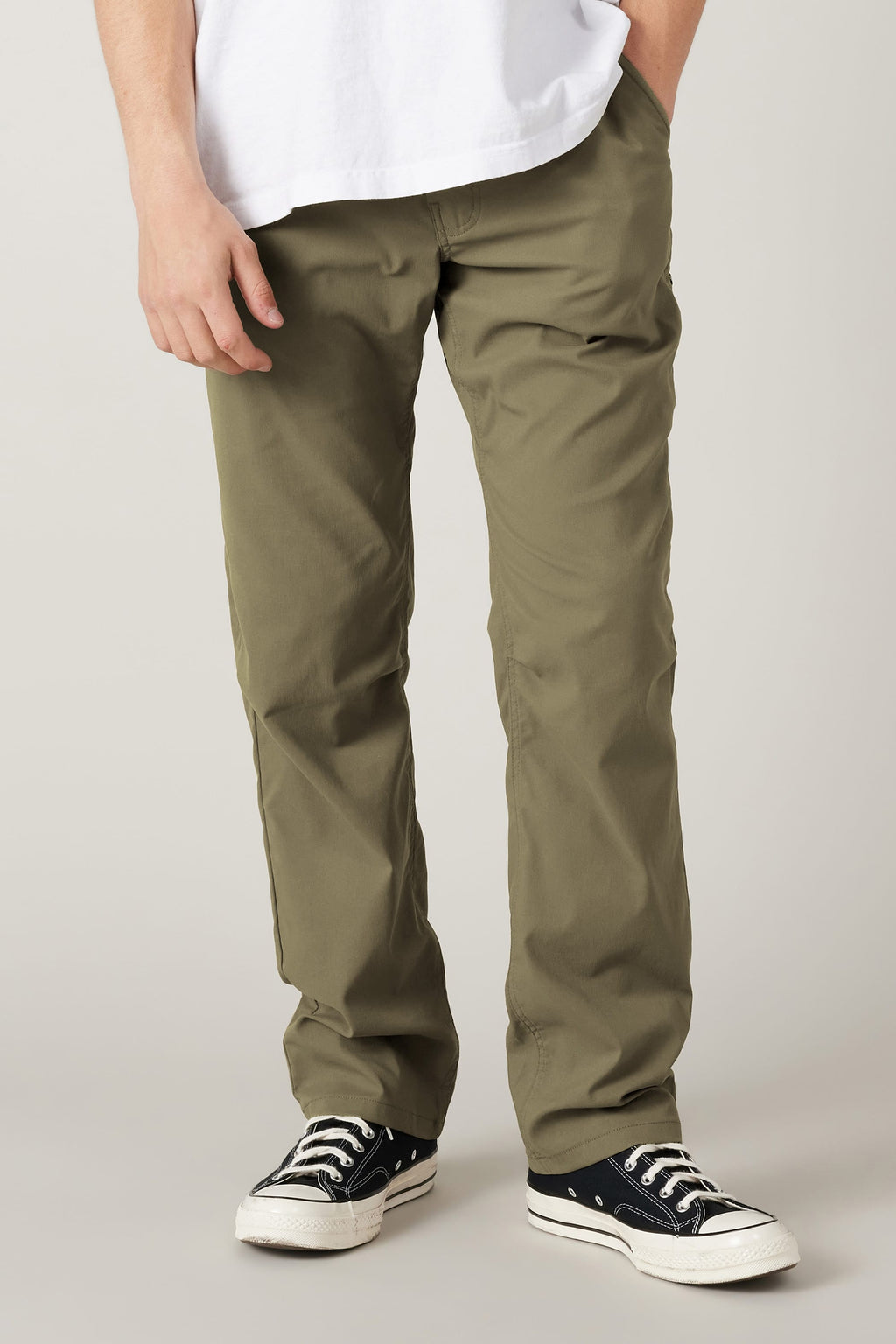 A person wearing the 686 Men's Everywhere® Pant - Relaxed Fit in olive, a white t-shirt, and black low-top sneakers with white soles stands against a plain background, one hand in their pocket.